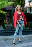 magnifique blond femme dans des lunettes habillé dans rouge veste et bleu jeans posant sur rue dans le ville photo