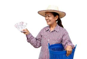 content Sénior femme agriculteur porte chapeau, plaid chemise, détient thaïlandais billets de banque argent et bleu panier, isolé sur blanc Contexte. concept, agriculteur content à avoir profit, le revenu de Faire agriculture. photo