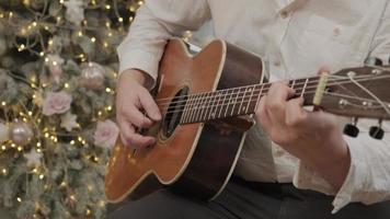 l'homme joue une chanson de noël à la guitare acoustique assis sur le sol près d'un arbre du nouvel an décoré dans des guirlandes lumineuses. une famille heureuse célèbre le réveillon de noël. fermer