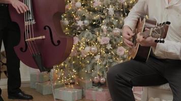 deux hommes jouent une chanson de noël à la guitare acoustique et au violoncelle près d'un arbre du nouvel an décoré dans des guirlandes lumineuses. une famille heureuse célèbre le réveillon de noël. photo