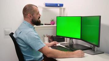 un officier masculin travaille sur un ordinateur avec deux maquettes d'écran vert à la maison. les employés sont assis devant des écrans.