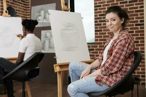 content Jeune femme séance à chevalet dans salle de cours et souriant à caméra, profiter amusement dessin classe avec amis, apprentissage esquisse techniques à groupe art classe. agréable Créatif loisir photo