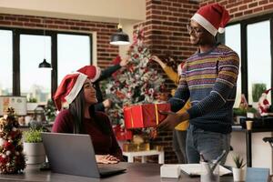 souriant africain américain Bureau ouvrier donnant Cristmas présent à asiatique femme collègue dans décoré bureau. diverse des employés partage Noël cadeaux tandis que travail sur portable dans espace de travail photo