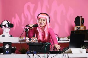 artiste avec rose cheveux mélange techno chanson avec électronique en utilisant professionnel platines tandis que profiter parlant avec Ventilateurs, ayant amusement ensemble pendant nuit temps dans club. femme profiter performant la musique photo