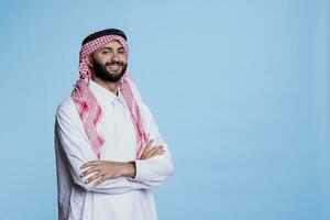 souriant musulman homme dans islamique traditionnel vêtements peignoir permanent avec plié mains et à la recherche à caméra avec de bonne humeur expression. insouciant arabe posant avec bras franchi portrait photo