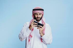 musulman homme portant traditionnel vêtements en jouant en ligne Jeu sur téléphone intelligent tandis que posant dans studio. arabe joueur dans thobe et islamique foulard profiter divertissement app sur mobile téléphone photo