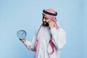 homme portant traditionnel musulman vêtements en portant rétro alarme l'horloge et à la recherche à temps. arabe la personne habillé dans thobe et foulard montrant ponctualité et programme date limite compte à rebours concept photo