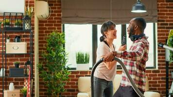 diverse couple ayant amusement nettoyage vivant pièce avec vide nettoyeur et serpillière, en chantant et écoute à musique. Jeune content les partenaires profiter printemps nettoyage, passe l'aspirateur appartement étages. photo