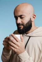 détendu arabe homme en portant blanc tasse dans mains tandis que profiter café. Jeune réfléchi la personne portant décontractée sweat à capuche vêtements en buvant chaud thé boisson de agresser sur bleu Contexte photo