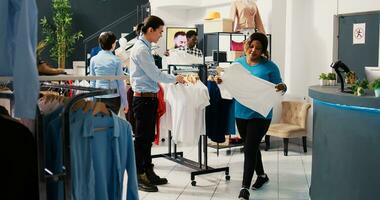 client nécessaire Aidez-moi avec branché chemises, discuter marchandise Matériel avec asiatique employé dans moderne boutique. africain américain femme achats pour élégant vêtements dans Vêtements boutique photo