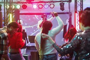 dj Parlant à public dans microphone tandis que performant sur étape dans boîte de nuit. gens dansant et élevage mains sur piste de dance tandis que assister électronique la musique concert dans club photo