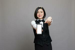 souriant asiatique serveuse La publicité téléphone intelligent et montrer du doigt à vide écran pour café menu promotion. de bonne humeur restauration un service femme employé dans uniforme en présentant mobile téléphone Vide écran tactile photo