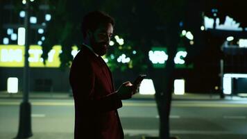 élégant homme en marchant autour ville à nuit, ambulant près Urbain grand bâtiment avec lumières. entrepreneur des promenades près grattes ciels dans ville centre illuminé par lampadaires, envoyer des SMS messages sur téléphone. photo