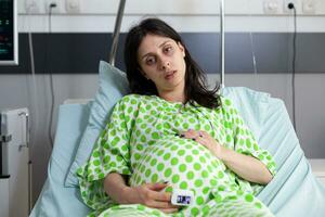 Enceinte patient portant médical oxymètre, à la recherche à caméra tandis que attendre à recevoir médical examen de médecin. femme attendant enfant et en train de préparer à donner naissance dans hôpital maternité pièce photo