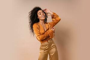 heureux africain femme avec parfait frisé Cheveux dans décontractée Orange chemisier et d'or un pantalon posant sur beige Contexte. photo