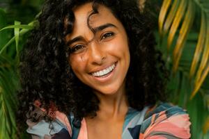 Extérieur portrait de souriant charmant femme avec frisé Cheveux posant plus de tropical des arbres et paume feuilles. photo