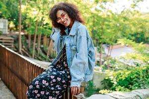 souriant femme avec afro coiffure séance sur une clôture dans été ensoleillé parc . portant jeans veste, robe avec floral impression et baskets . photo