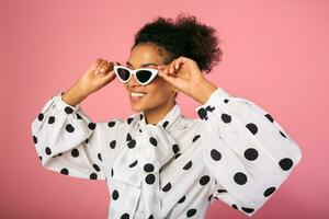 studio photo de africain noir femme dans élégant robe et blanc des lunettes de soleil. jolie souriant femelle posant plus de rose Contexte.