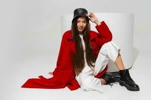 à la mode modèle dans élégant chapeau, rouge manteau et bottes posant sur blanc Contexte dans studio. photo