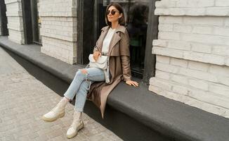 élégant à la mode femme avec court brune Cheveux portant branché des lunettes de soleil, beige Couleur manteau, blanc texturé cuir épaule sac, en marchant dans rue de européen ville. photo