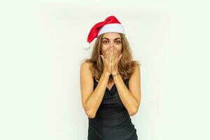 jolie femme avec surprise visage dans Père Noël chapeau et relégant noir robe posant sur blanc Contexte dans studio. photo