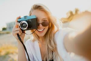 svelte blond femme en portant rétro caméra et ayant amusement sur ensoleillé plage. Naturel beauté. photo