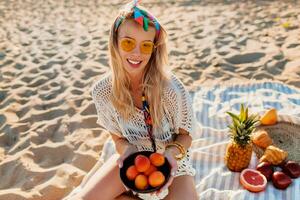 magnifique blond fille en portant assiette avec pêche et relaxant sur le soir tropical plage, le coucher du soleil couleurs. séance sur serviette. tropical des fruits. portant élégant Bandeau et boho robe. en bonne santé vie. photo