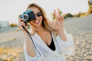 blond femme en portant rétro caméra et ayant amusement sur chaud ensoleillé plage. des lunettes de soleil, blanc tenue. photo