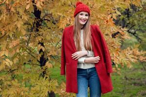 l'automne mode portrait de content blond femme dans rouge élégant manteau et tricoté chapeau en marchant dans parc. photo