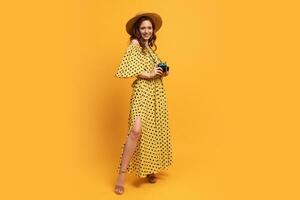 plein longueur photo de en voyageant femme dans paille chapeau et été robe posant avec rétro caméra plus de Jaune backgrond dans studio.