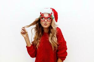 joyeux insouciant blond femme dans mignonne mascarade des lunettes et Nouveau année chapeau dans rouge tricoté chandail posant sur blanc Contexte . isoler. Noël et Nouveau tester fête concept. photo