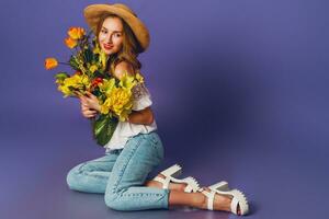 proche en haut printemps portrait de magnifique blond Jeune Dame dans élégant paille été chapeau en portant coloré printemps fleur bouquet près violet mur Contexte. photo