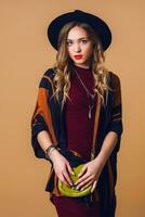 studio portrait de Jeune Frais blond femme dans marron paille poncho , la laine noir branché chapeau et rond des lunettes à la recherche à caméra. vert cuir avait sac. photo