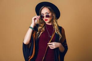 studio proche en haut portrait de Jeune Frais blond femme dans marron paille poncho , la laine noir branché chapeau et rond des lunettes à la recherche à caméra. vert cuir avait sac. photo