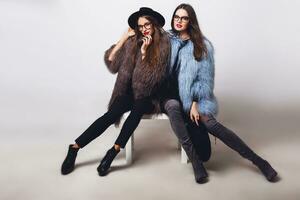 mode portrait de deux élégant femmes dans élégant hiver décontractée tenue, branché lunettes, brillant faire en haut, rouge lèvres séance sur blanc tableau. plein longueur . photo