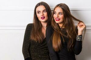 deux Jeune élégant souriant femmes, copains dans soir décontractée tenue ayant amusement sur blanc Urbain mur. brillant faire en haut. photo