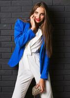 à la mode studio image de élégant attrayant Jeune femme dans décontractée blanc costume et bleu veste en portant luxe bourse posant sur noir Urbain brique mur Contexte. photo