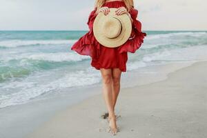 blanc gingembre femme dans élégant robe en portant paille chapeau et posant sur le plage près océan. blu ciel. venteux temps photo