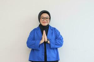 magnifique Jeune asiatique femme dans lunettes, hijab et portant bleu blazer posant namaste salutation mains avec souriant visage photo