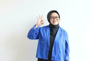 magnifique Jeune asiatique musulman femme, portant des lunettes et bleu blazer avec main geste de approbation ou D'accord photo