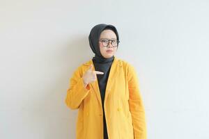 magnifique Jeune asiatique musulman femme dans lunettes, hijab et portant Jaune blazer avec main montrer du doigt de côté photo