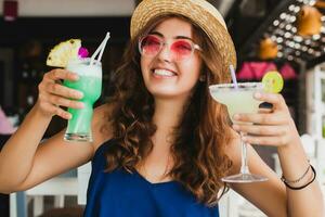 attrayant Jeune femme dans bleu robe et paille chapeau portant rose des lunettes de soleil en buvant des cocktails photo