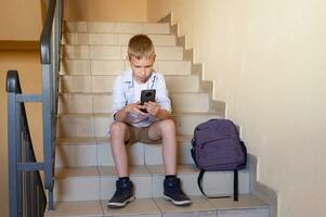 une adolescent garçon est à la recherche à le sien téléphone tandis que séance sur le pas avec une sac à dos. photo