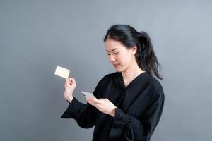 portrait d'une jeune fille asiatique heureuse montrant une carte de crédit en plastique tout en tenant un téléphone portable sur fond gris photo