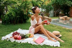 gracieux asiatique femme profiter été pique-nique sur vert pelouse près bassin. portant romantique rose robe, en train de lire livre. Frais des fruits et des croissants. paille chapeau. photo