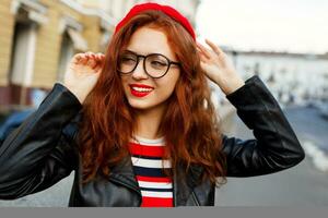 élégant Jeune gingembre femme dans rouge béret et des lunettes posant sur le rue. photo