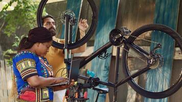 vibrant africain américain Dame navigation sur l'Internet pour vélo équipement entretien. noir femme vérification pour réparation instructions sur portable tandis que caucasien homme démantèle cassé bicyclette roue. photo