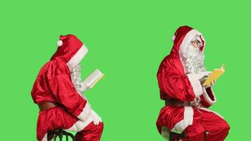 sage Père Noël claus en train de lire livre plus de écran vert toile de fond, profiter poésie ou Littérature livre séance sur chaise. sur de soi homme habillé comme père Noël avec conférence passe-temps, connaissance. photo