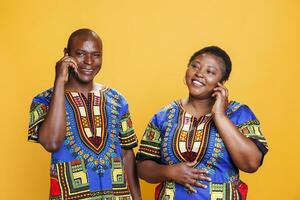 de bonne humeur africain américain couple portant ethnique vêtements bavardage sur téléphone intelligent. souriant mature homme et femme répondre téléphone intelligent appel et ayant amusement la communication portrait photo