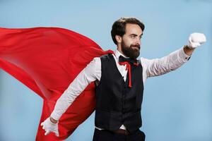 déterminé adulte posant comme super-héros tandis que il porte rouge manteau contre bleu Contexte dans studio, professionnel la personne agissant comme une surhumain. homme avec formel tenue et dessin animé cap. photo